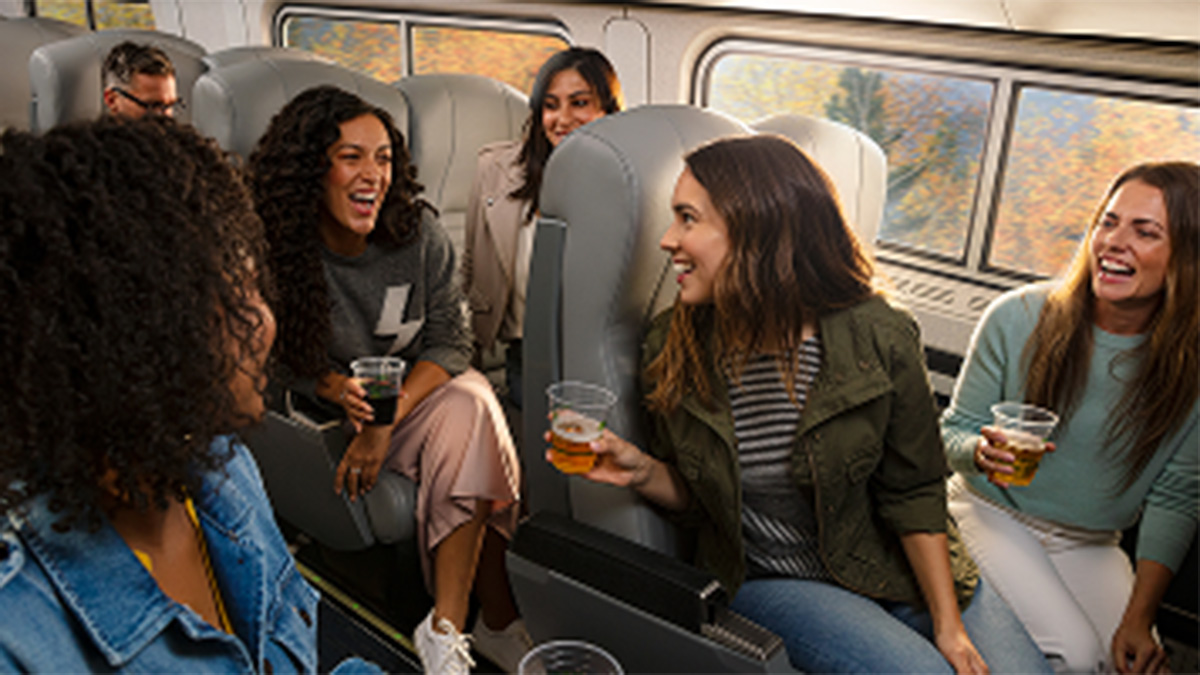 A group of diverse women laugh and talk with drinks in a train car