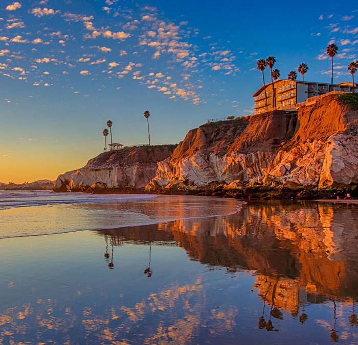 A scenic beach featuring a cliffside with a house perched atop, overlooking the reflective ocean below.